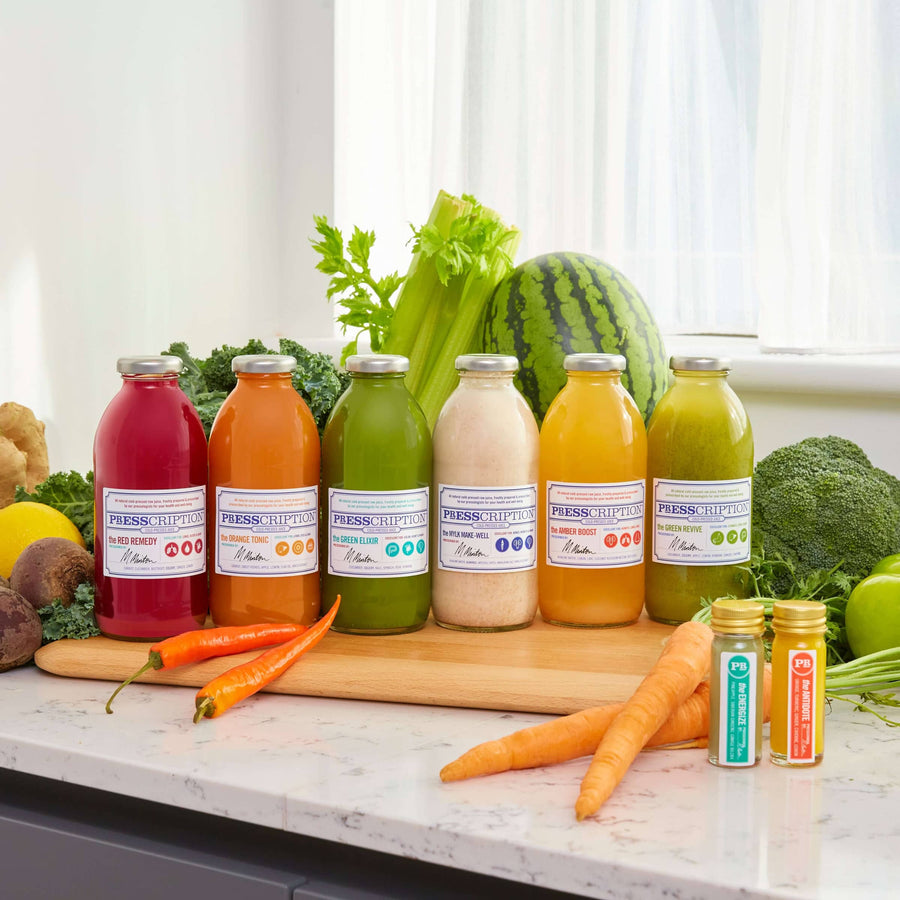 Presscription cold-pressed juices and shots on a kitchen counter with fresh fruit and vegetables
